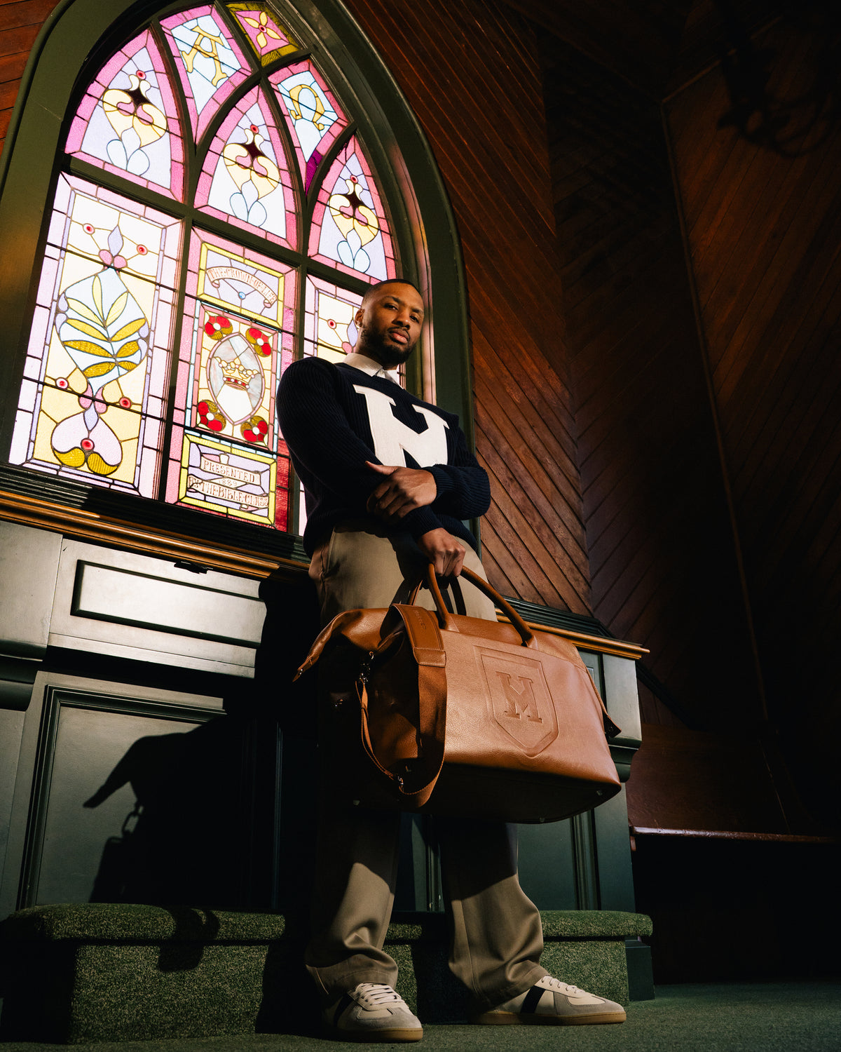 Damian Lillard stands in a dimly lit church in front of a stained-glass window, holding a the Honorable Mention vegan leather weekender. He wears a dark sweater with a large white logo on the chest, beige pants, and white sneakers.