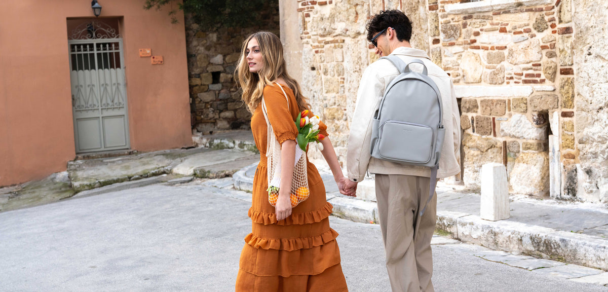 A woman in a rust-orange tiered dress holding hands with a man wearing a cream shirt, beige trousers, and carrying the Nostos Matte backpack in Pebble grey, walking down a stone street with a small bouquet of flowers.