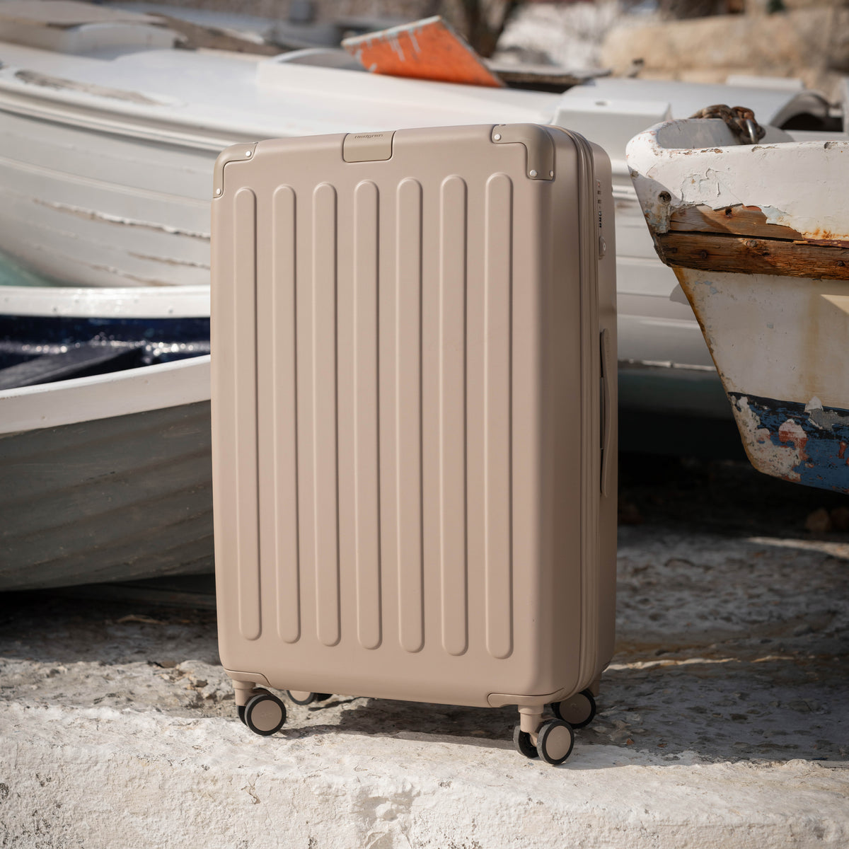 The Nostos Medium Matte Spinner in Sahara, standing on a concrete surface with weathered boats in the background.