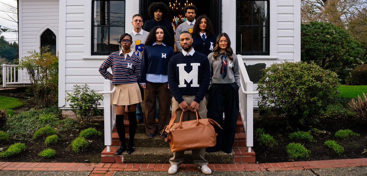 A group of nine people posing together on the front steps of a white house. Damian Lillard is in the front wearing a navy sweater with a large white letter 'M' and holding the Honorable Mention Weekender.