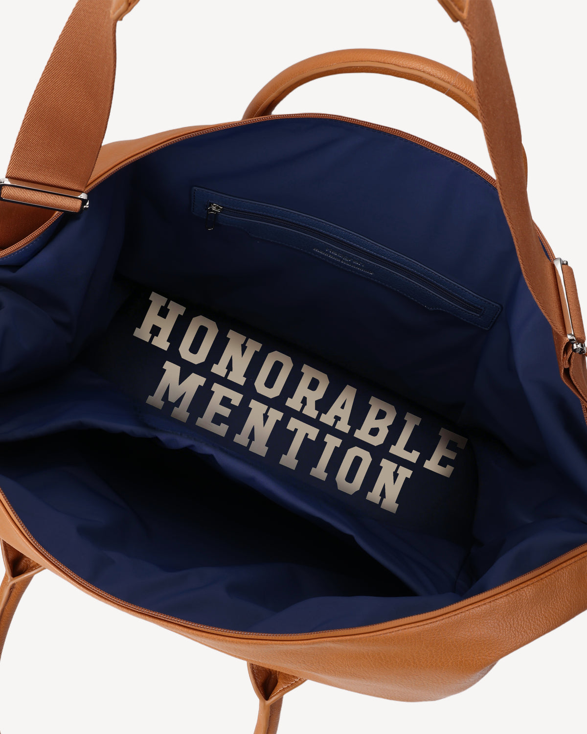 Close-up of the interior of the Honorable Mention Weekender in Tuscany vegan leather with a navy blue lining. The lining features the text 'HONORABLE MENTION' printed in large white block letters, and a zippered pocket is visible at the top.